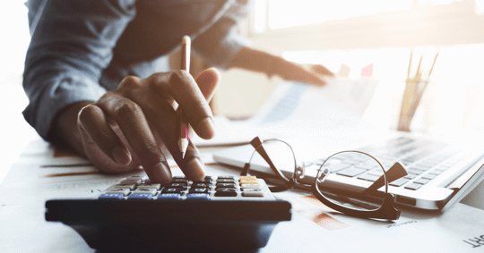 Someone types information into a calculator near a laptop and glasses