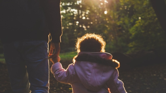 Man holds hand with young child in puffy jacket, walking away from the camera