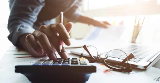 Person types into a calculator next to glasses and a laptop